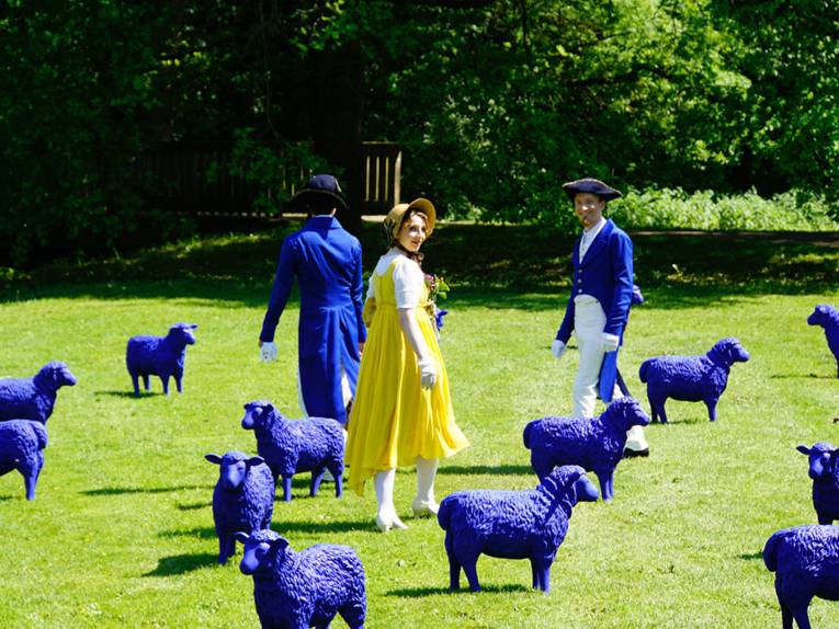 Historisch gekleidete Männer und Frauen in einer Parkanlage mit blauen Schafsmodellen