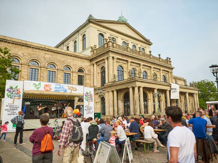 Besucherinnen und Besucher des Autofreien Sonntags an Biertischgarnituren auf dem Opernplatz schauen sich das Programm auf der Bühne von "Landwirt schafft Zukunft" an