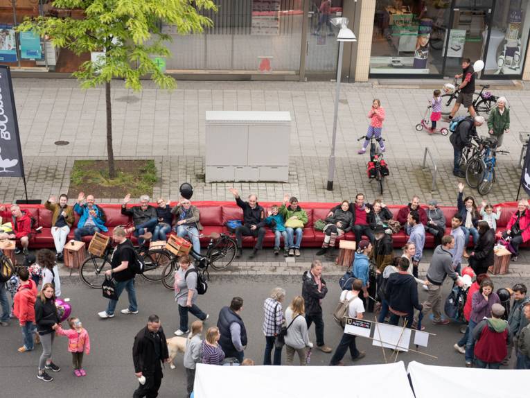 Auf einem überlangen Sofa, das am Straßenrand aufgebaut ist, sitzen achtzehn Personen - zum Teil winkend - und schauen den Fußgängern beim Flanieren zu.