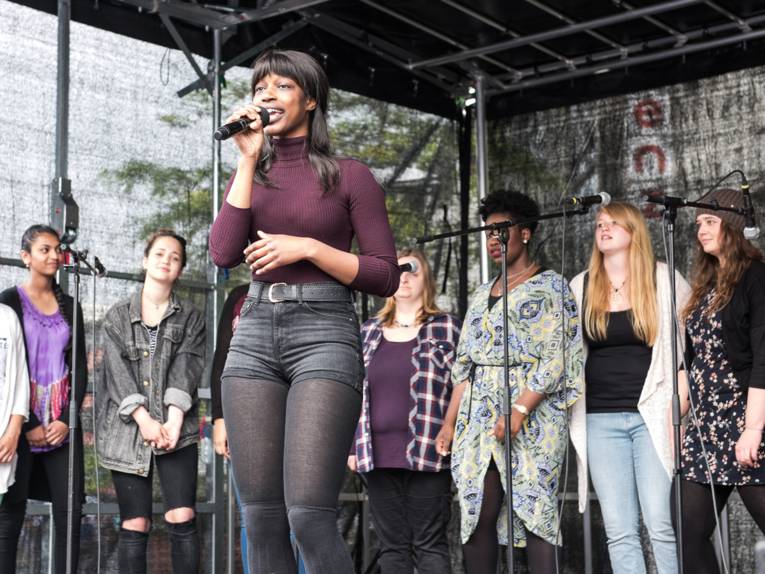 Eine Sängerin mit Mikrophon im Vordergrund und acht Background-Sängerinnen auf einer Bühne