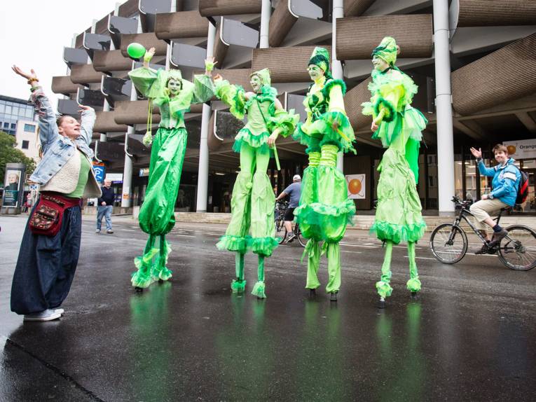 Vier grün gekleidete Schauspielerinnen auf Stelzen mit einem im Vorbeifahren winkenden Radfahrer und einer fröhlichen Besucherin im Vordergrund.