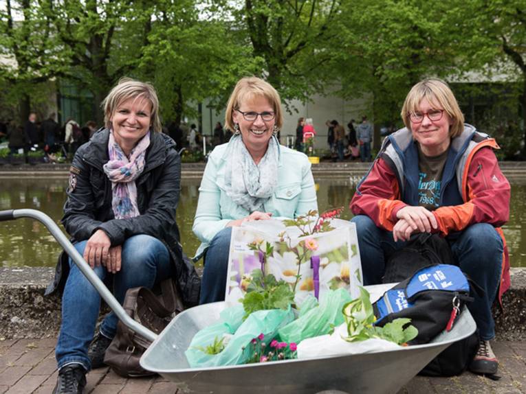 Drei Besucherinnen sitzen auf einer Teichumrandung und haben vor sich eine mit Pflanzeneinkäufen gefüllte Karre