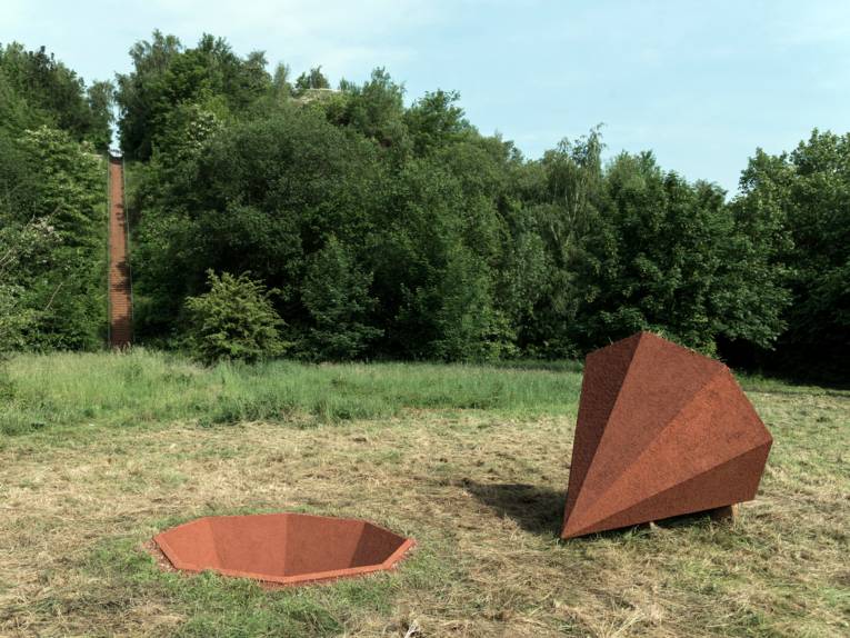 Aus dem grasbewachsenen Boden scheint ein achteckiger Kegel ausgestochen zu sein, der neben dem Loch liegt.
