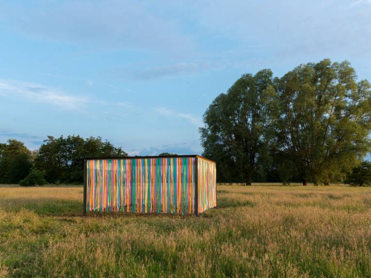 Eisenträger bilden ein Rechteck, an den Seiten hängen bunte Kunststoffstreifen. Der Wind bewegt die Kunststoffstreifen.