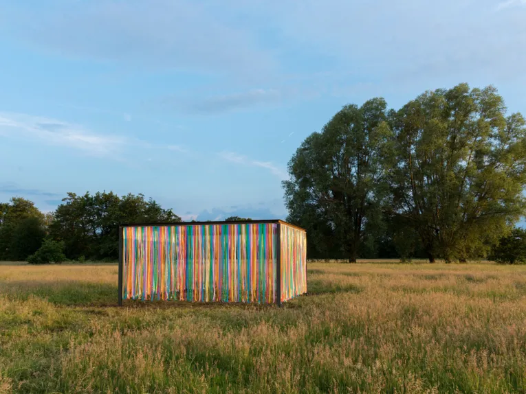 Eisenträger bilden ein Rechteck, an den Seiten hängen bunte Kunststoffstreifen. Der Wind bewegt die Kunststoffstreifen.