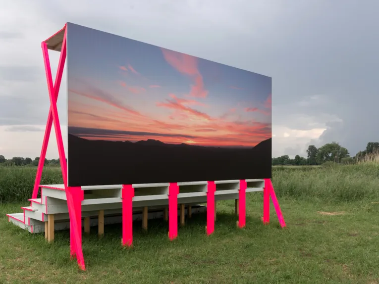 Eine überdachte Tribüne gibt den Blick auf die Landschaft frei. Die Rückseite der Tribüne zeigt einen Sonnenuntergang, der sich unter optimalen Bedingungen vielleicht in der Landschaft wiederfinden lässt.