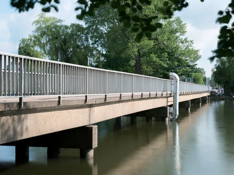 An der Fußgängerbrücke zur Badeinsel ist eine silbrige Röhre angebracht. Die Röhre funktioniert wie ein Periskop, wer hindurchschaut, blickt in einen gestalteten Raum unter der Brücke.