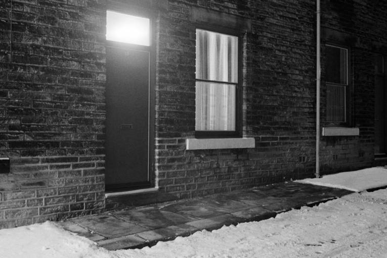 Schwarzweißfoto einer Häuserzeile im Winter, bei der nur vor einem Haus der Gehweg vom Schnee geräumt wurde.