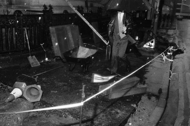 Schwarzweißfoto eines Mannes, der bei starken Regen mit Regenschirm in eine Baustelle auf einem Fußweg geht.