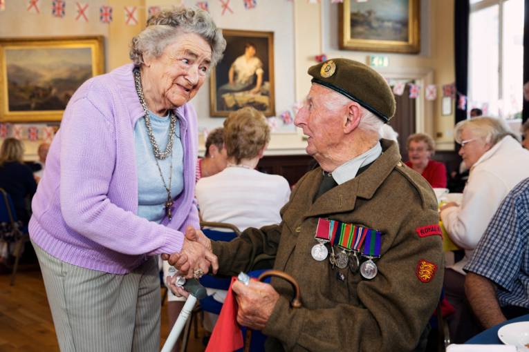 Alte Frau gibt einem alten Mann in ordenbehängter Uniform die Hand.