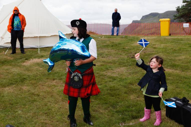 Junges Mädchen in Kilt mit aufblasbaren Delfin in den Händen, daneben kleines Mädchen mit schottischen Fähnchen in den Händen.