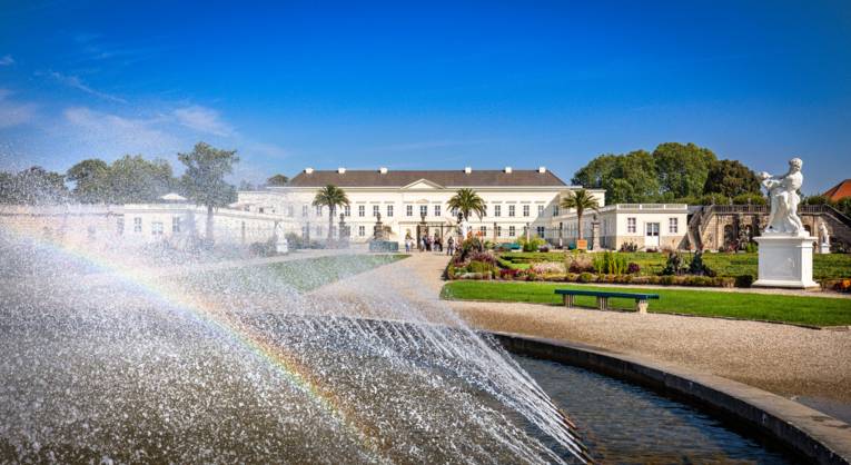 Die Sonne zaubert einen kleinen Regenbogen in den Springbrunnen im Großen Garten. Im Hintergrund ist das Schloss Herrenhausen zu sehen.