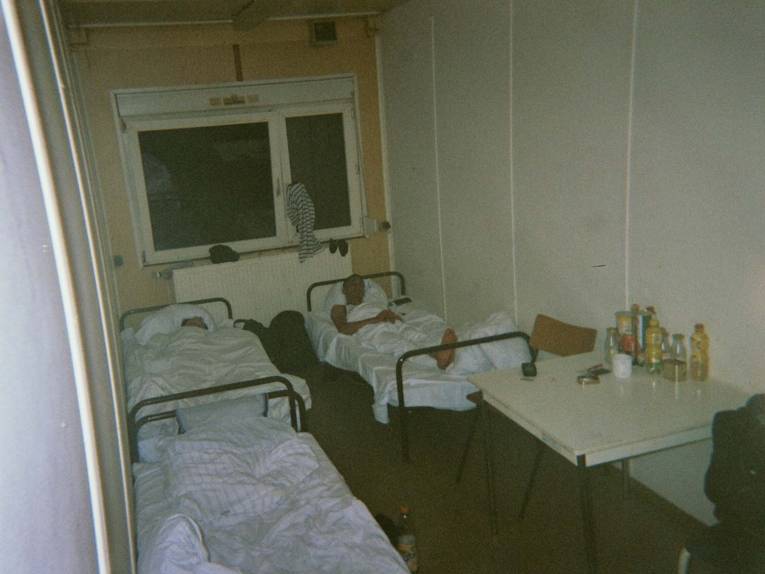 Fotografie, die zwei Männer in einem dunklen Container zeigt. Jeder liegt in seinem Bett. 
