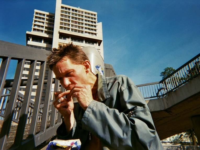 Foto, das einen Mann zeigt, der sich auf einem Platz eine Zigarette dreht. 