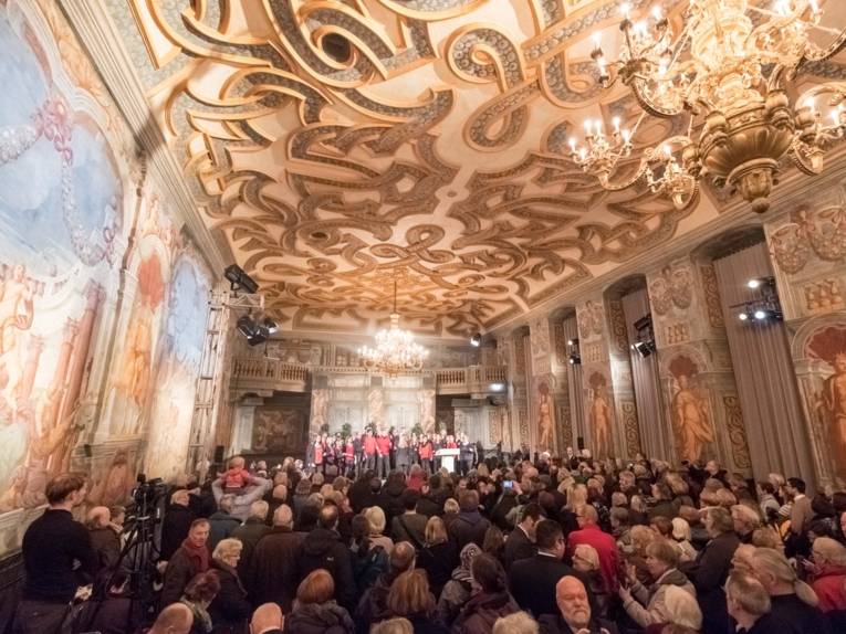 Chor auf einer Bühne in einer Galerie. Davor viele Menschen. 