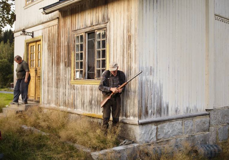 Alter Mann mit einem Gewehr und alte Frau vor einem Holzhaus.