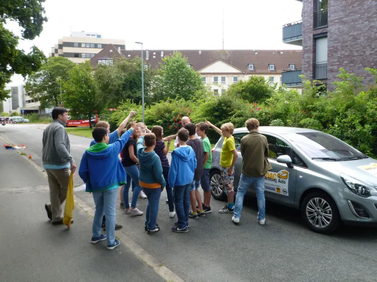 Sch&uuml;lerinnen und Sch&uuml;ler umringen einen Trainer auf der Stra&szlig;e, der neben einem Auto steht und eine Frage gestellt hat.