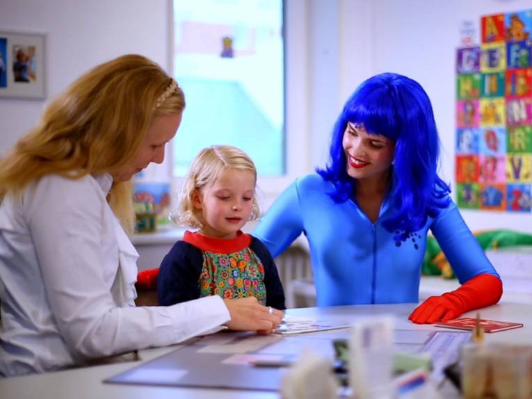 Zwei Frauen und ein Kind sitzen an einem Tisch. Die Frau links trägt einen weißen Arztkittel, die Frau rechts ein blaues Superheldinnengewand.