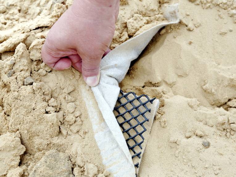 Eine Hand schiebt ein Vlies zur Seite, darunter sind eine gitterartige Matte und eine Kunststoffabdichtung zu sehen. Der ganze Aufbau ist von sandigem Boden umgeben.
