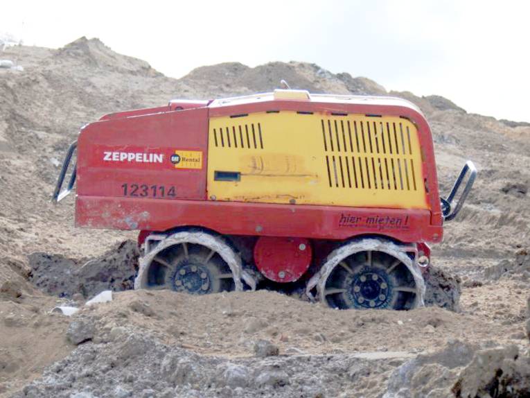 Ein Walzenfahrzeug in den Farben Gelb und Rot mit zwei Achsen fährt über unebenes Gelände.