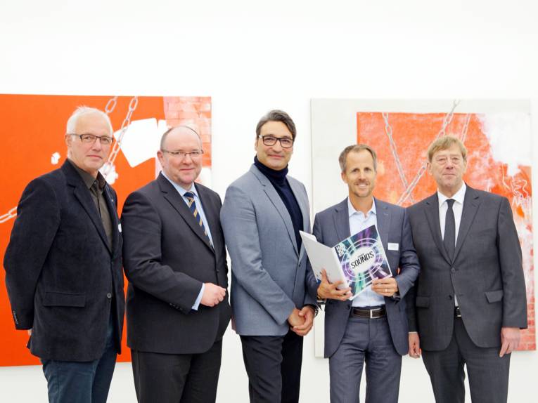 Fünf Männer stehen nebeneinander vor einer Wand mit abstrakter Malerei.