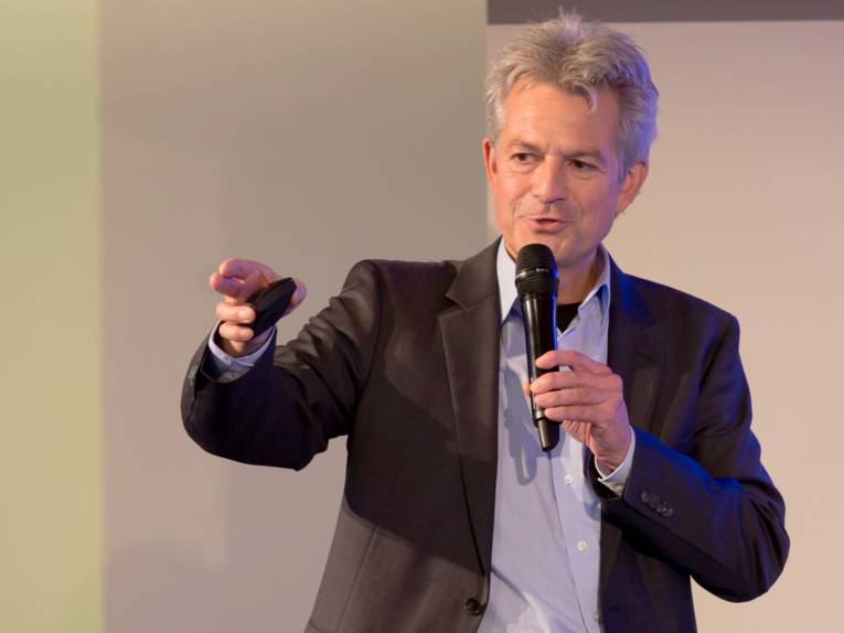 Ein Mann spricht in ein Mikrofon und macht mit der rechten Hand eine zeigende Bewegung. Rechts hält er eine Fernbedienung.