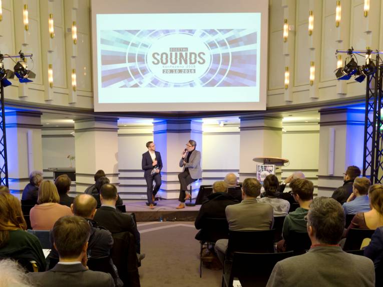 Das Publikum verfolgt ein Interview zwischen zwei Männern auf dem Podium.