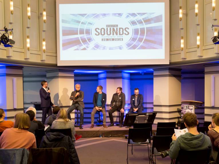 Gäste sitzen auf Stühlen und verfolgen, was auf dem Podium geschieht. Dort steht der Moderator und vier Männer sitzen nebeneinander.