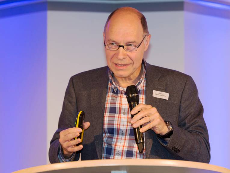 Ein Mann steht hinter einem Rednerpult, in den Händen hält er ein Mikrofon und eine Fernbedienung.