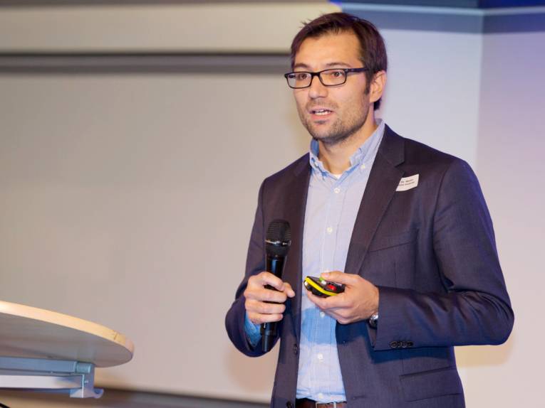 Ein Mann steht neben einem Rednerpult, in der einen Hand hält er ein Mikrofon, in der anderen eine Fernbedienung.