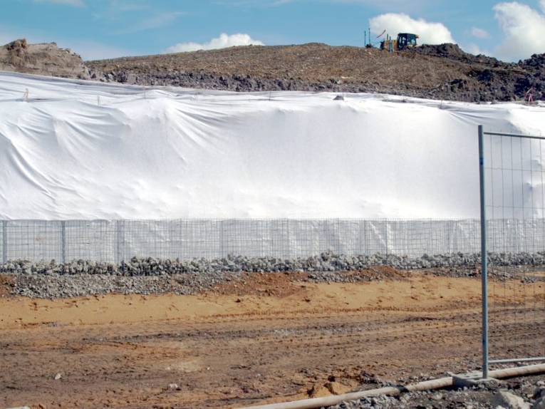 Blick auf die Fulgurithalde. Die Erhebung ist mit weißem Geovlies abgedeckt, davor stehen Drahtschotterkästen (Gabionen), die etwa zu einem Drittel mit Steinen gefüllt sind. Auf dem nicht abgedeckten Teil der Erhebung ist ein Baufahrzeug.