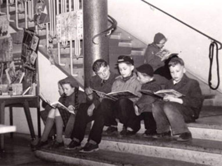 Schwarz-weiß Aufnahme von Kindern aus den 1960er Jahren, die lesend auf der Treppe im Haus der Jugend sitzen