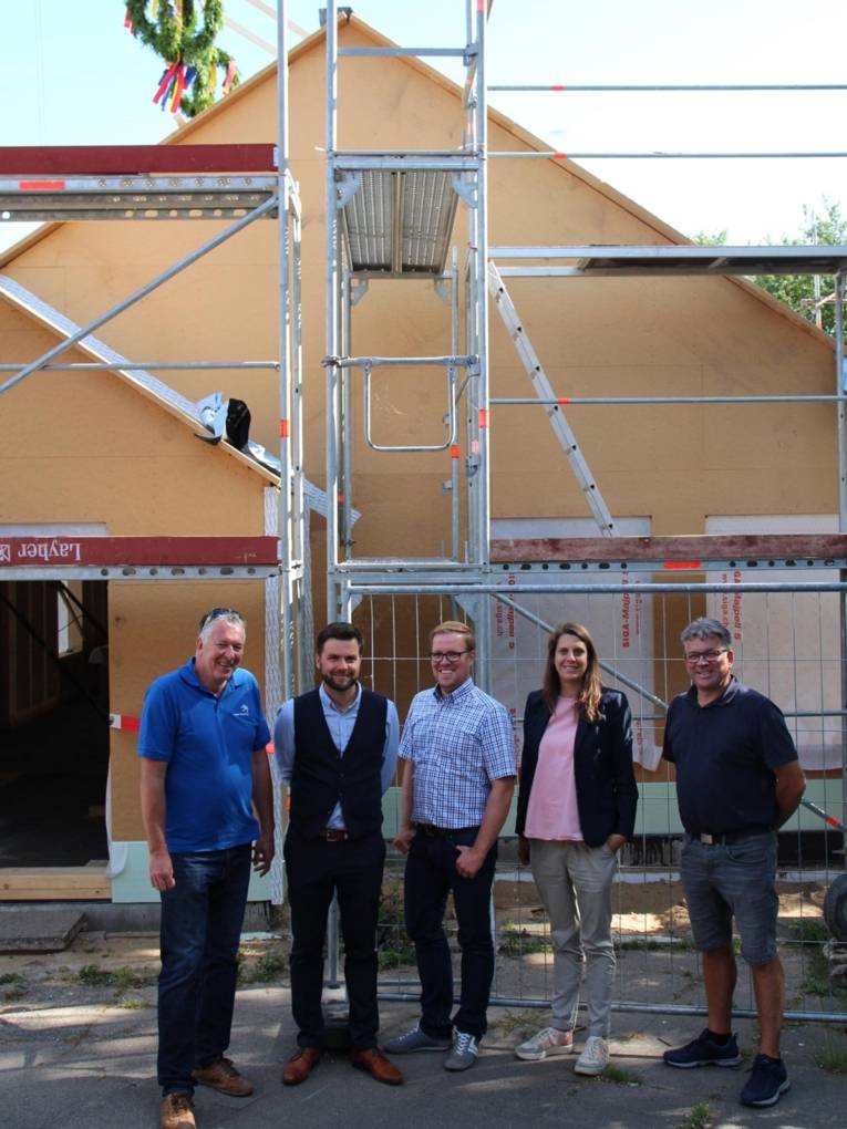 Eine Frau und vier Männer vor einem Gebäude mit Baugerüst und Richtkrone.