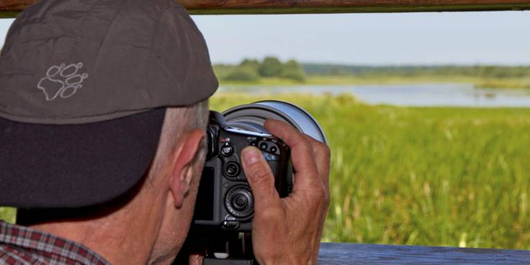 Ein Mann ist an einem Aussichtspunkt und schaut durch den Sucher seiner Kamera auf ein Feuchtgebiet.