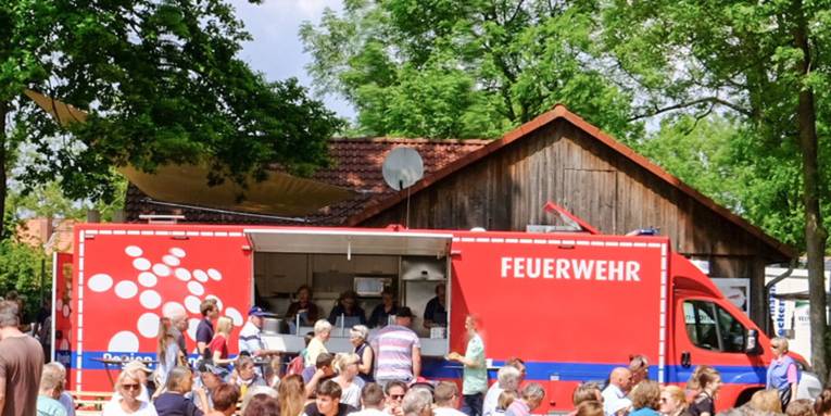 Mesnchen stehen schlange an am Gerätewagen Küche der Freiwilligen Feuerwehr. Dafür spielen sich Szenen eines Dorffestes ab.