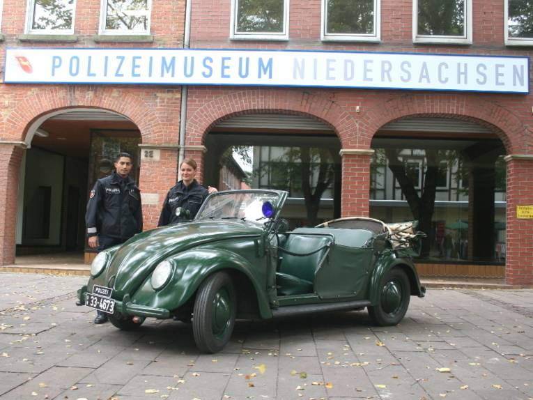 Eine Polizistin und ein Polizist stehen neben einem Polizeieinsatzfahrzeug VW Käfer vor dem Polizeimuseum Niedersachsen.