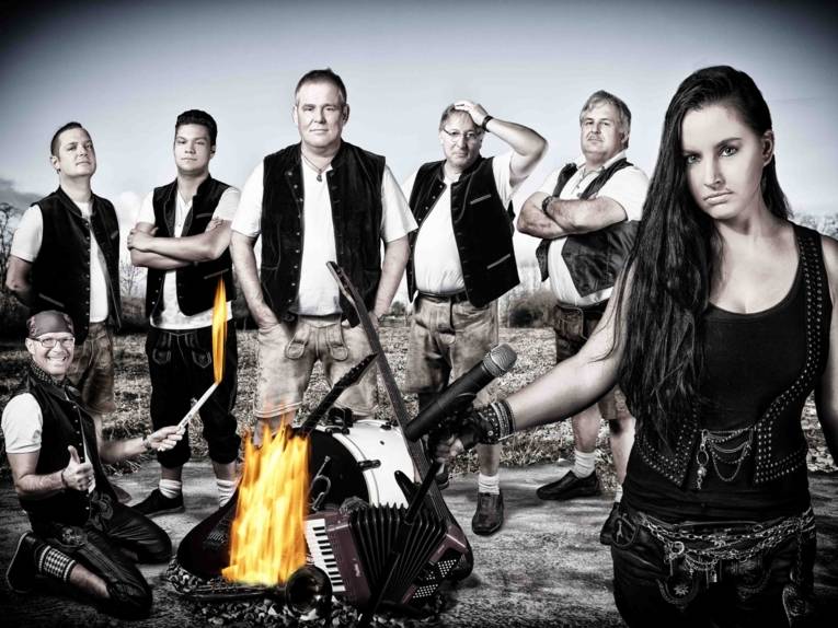 Sechs Männer und eine Frau mit Instrumenten um ein Feuer. 