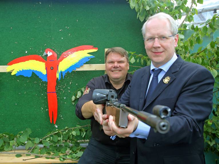 Zwei Männer mit einem Gewehr. Im Hintergrund ein Holzpapagei. 