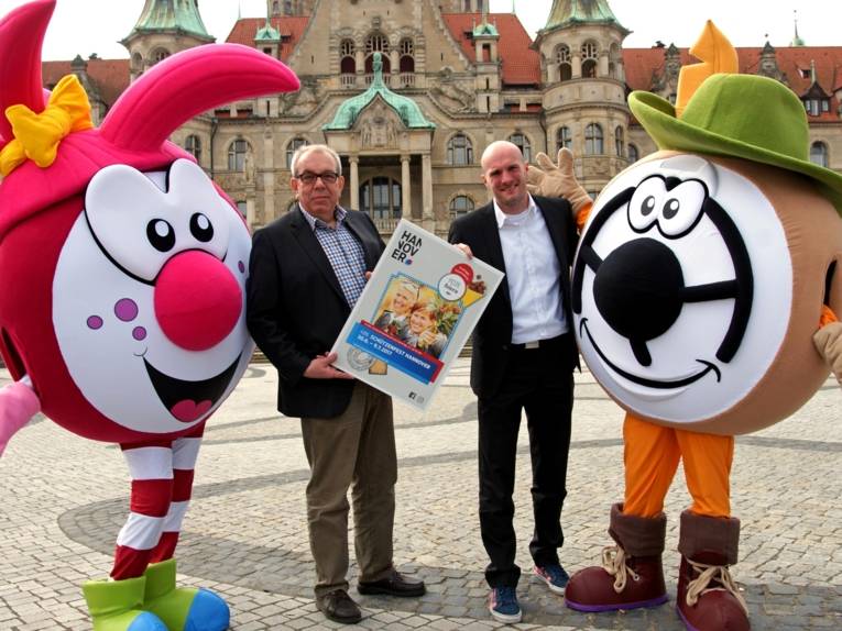 Zwei Männer mit zwei Maskottchen vor einem Rathaus.