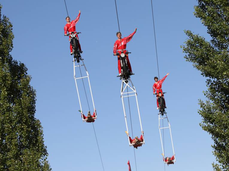 Motorradfahrer, die über ein Seil durch die Luft fahren. Unter den Fahrzeugen schaukeln Artisten. 