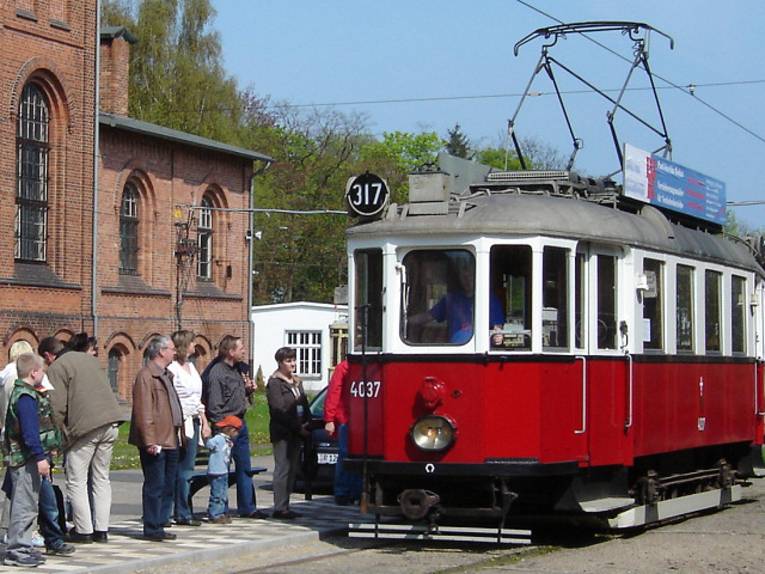 Historischer Straßenbahnwagen an einer Haltestelle. Personen stehen an um einzusteigen.