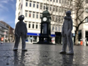 Zwei Lehmfiguren, die vor einer Innenstadt-Uhr stehen. Durch die Perspektive, sehen die Figuren menschengroß aus
