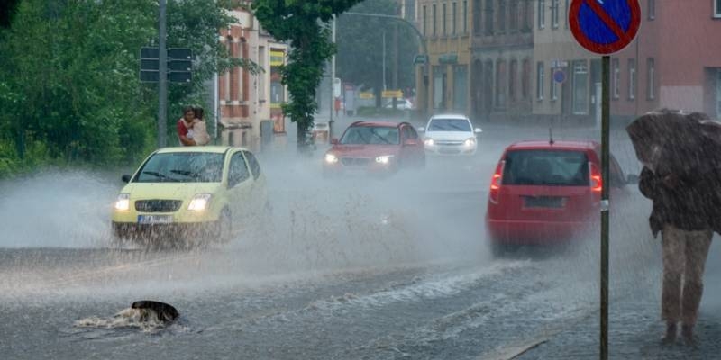 Vier Autos bei starkem Regen auf einer Straße, Wasser drückt einen Gulli hoch, ein Schild und ein Fußgänger mit Schirm.
