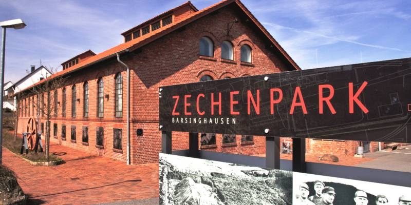 Schild mit der Aufschrift "Zechenpark. Barsinghausen", dahinter ist ein Backsteingebäude.
