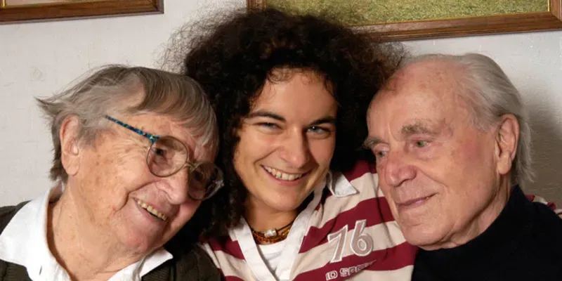 Eine junge Frau, die rechts im Arm eine Seniorin h&auml;lt und links im Arm einen Senior. Hinten an der Wand sind Teilst&uuml;cke zweier Gem&auml;lde zu sehen.