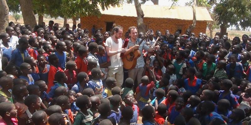 Zwei Musiker treten zwischen Schülerinnen und Schülern aus Malawi unter freiem Himmel auf
