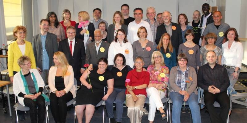 Etwa 25 Personen sitzen und stehen vor eine B&uuml;hne.