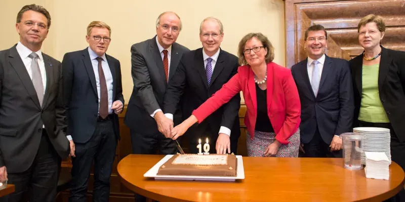 Zwei Frauen und f&uuml;nf M&auml;nner schneiden eine Torte an.