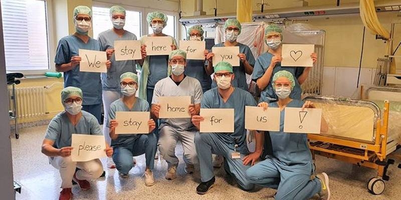 Medizinisches Personal mit Mundschutz, alle halten Kärtchen hoch deren Aufschriften zusammen den Satz "We stay here for you, please stay home for us" ergeben.