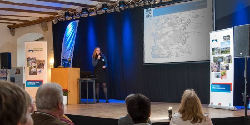 Blick auf eine Bühne, dort steht eine Frau an einem Rednerpult.